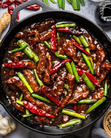 A cast iron skillet filled with saucy Mongolian beef strips, red chili peppers, and green onions, surrounded by ginger, garlic, dried red chilies, a bowl of sesame seeds, and a jar of soy sauce on a marble surface.