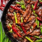 A cast iron skillet filled with spicy stir-fried Mongolian beef strips, red chili peppers, green onions, and sesame seeds in a glossy brown sauce. Fresh green onions and dried red chilies are visible around the skillet.