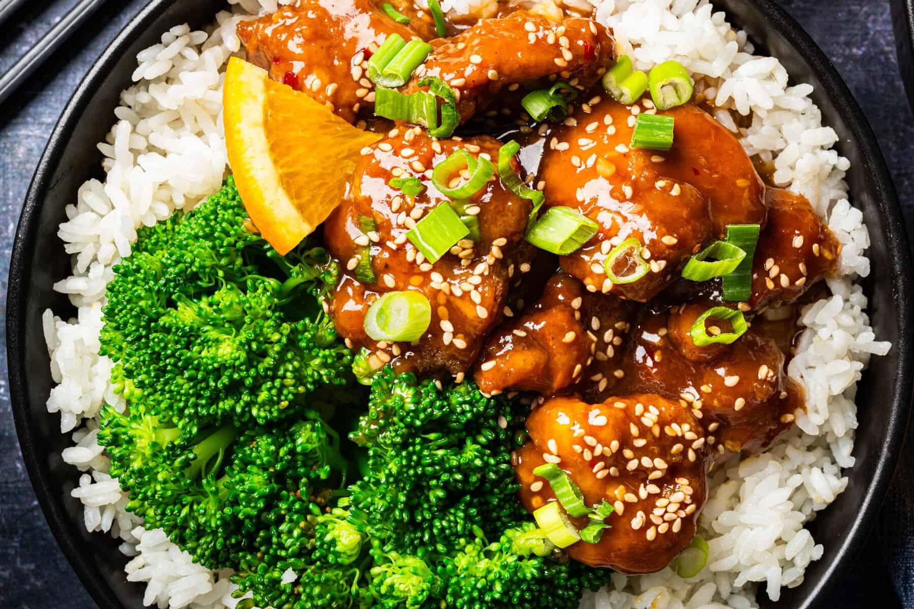 Orange Chicken (with white rice and broccoli) A bowl of white rice topped with broccoli florets, orange-glazed chicken garnished with sesame seeds and chopped green onions, with a slice of orange on the side.