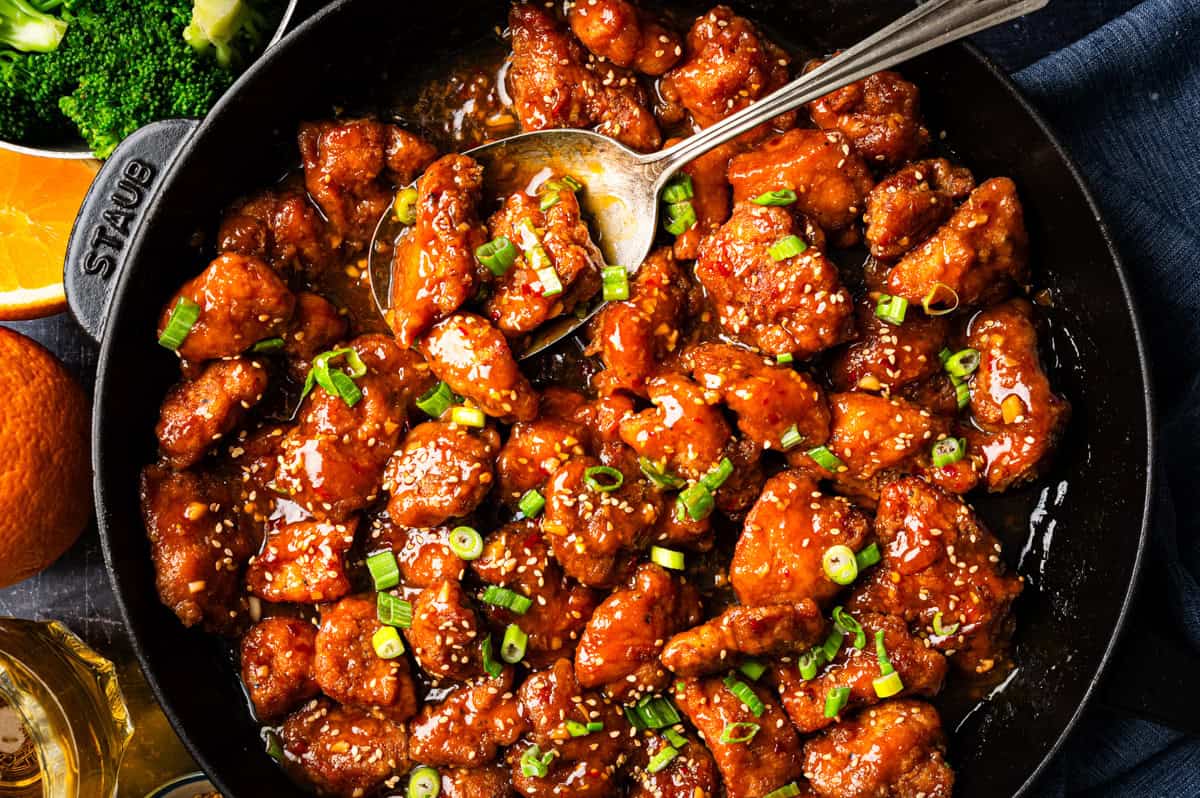 Orange Chicken A skillet filled with glazed orange chicken pieces garnished with chopped green onions and sesame seeds, with a metal spoon resting inside.