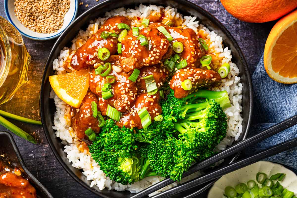 Orange Chicken (with white rice and broccoli) A bowl of white rice topped with glazed orange chicken, sesame seeds, chopped green onions, and broccoli, garnished with an orange slice. Chopsticks rest on the bowl and fresh oranges are in the background.