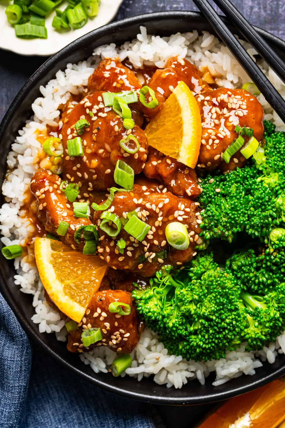 Orange Chicken (with white rice and broccoli) A bowl of white rice topped with orange chicken pieces, steamed broccoli, sliced green onions, sesame seeds, and orange wedges, with black chopsticks resting on the bowl's rim.