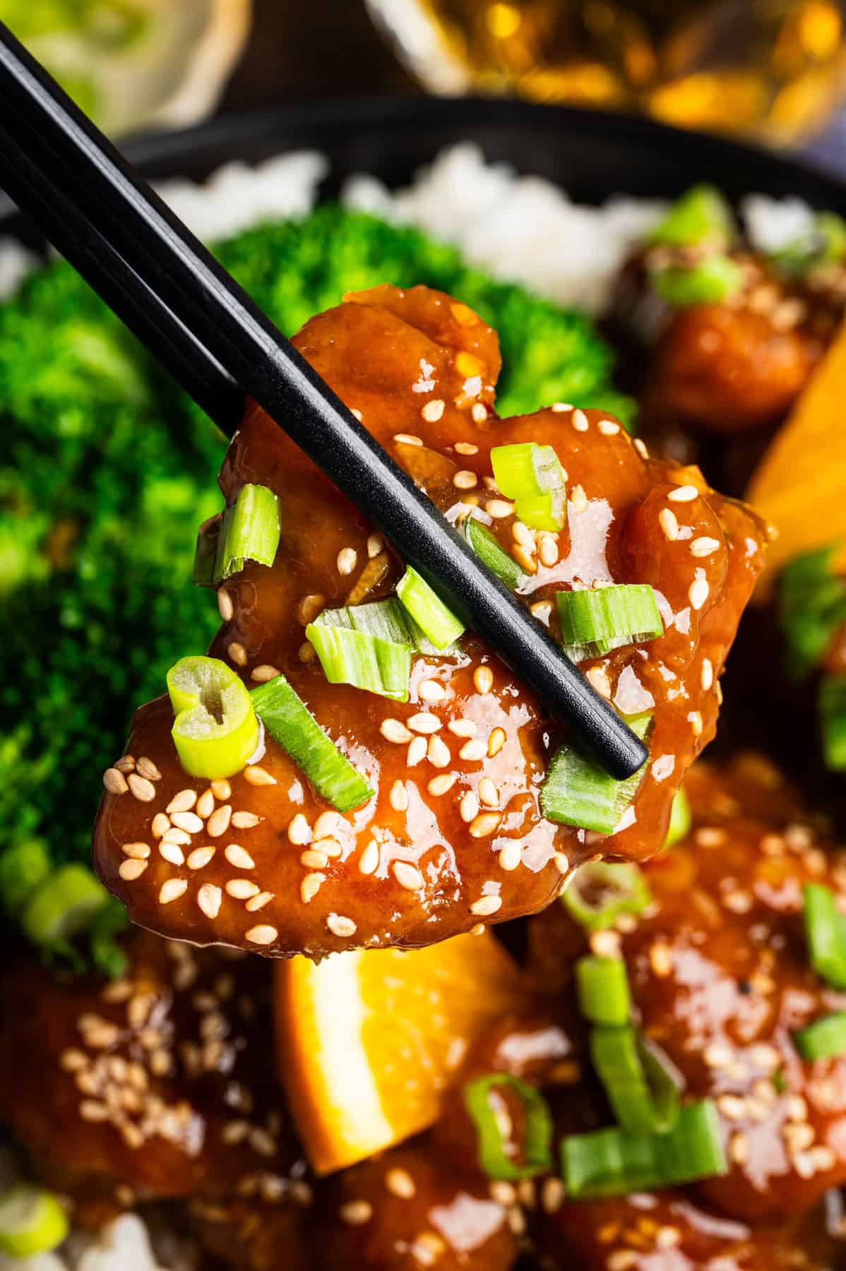 Orange Chicken Close-up of black chopsticks holding a piece of sesame-covered orange chicken garnished with chopped green onions, with broccoli and rice visible in the background.