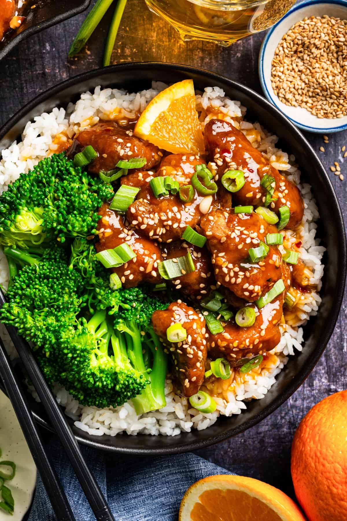 Orange Chicken (with white rice and broccoli) A bowl of white rice topped with glazed orange chicken, steamed broccoli, sliced green onions, sesame seeds, and a wedge of orange. Chopsticks rest on the bowl, and there are extra sesame seeds and oranges nearby.