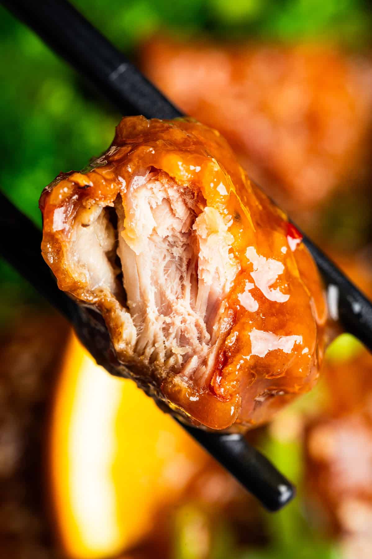 Orange Chicken A close-up of chopsticks holding a piece of crispy chicken coated in a glossy orange sauce, with a bite taken out showing the juicy interior.
