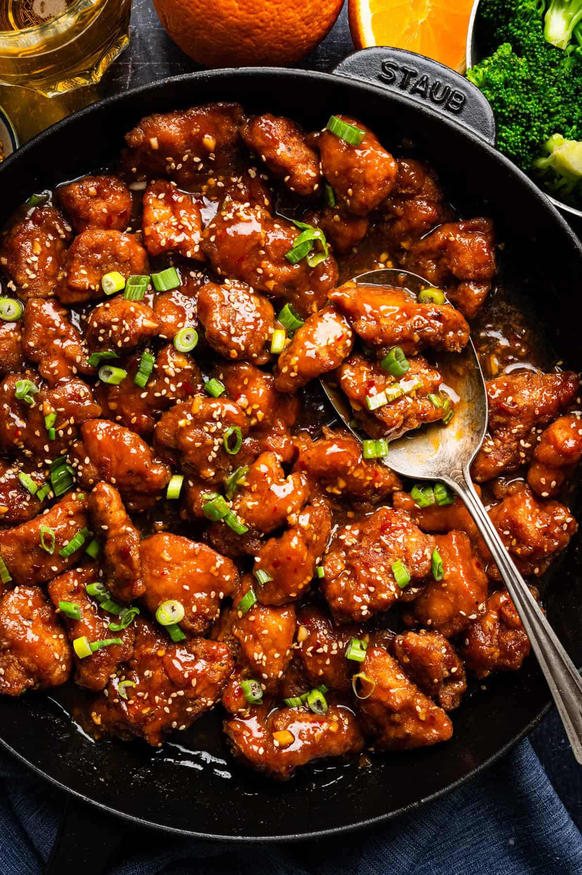 Orange Chicken A skillet filled with saucy, glazed orange chicken pieces garnished with chopped green onions and sesame seeds. A spoon rests in the skillet.