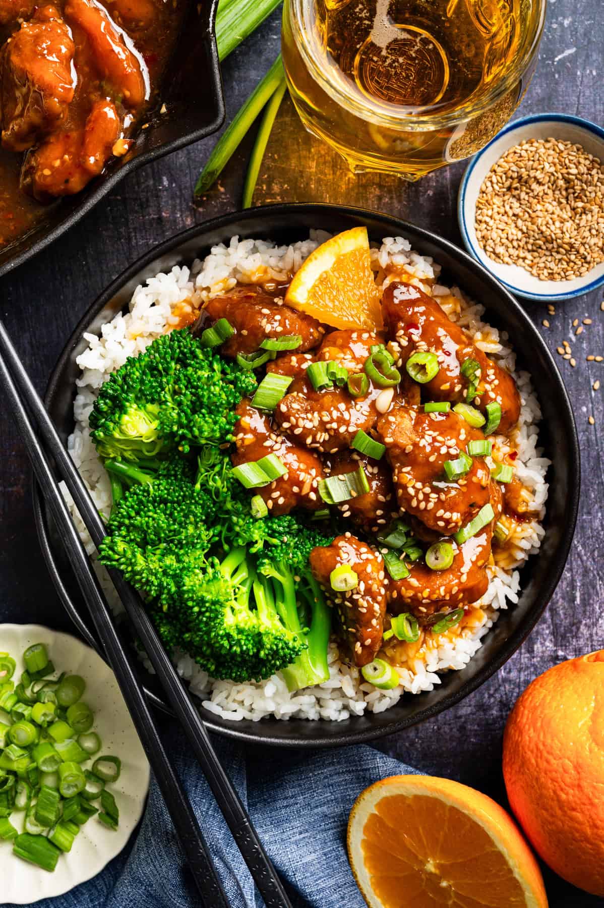 Orange Chicken (with white rice and broccoli) A bowl filled with white rice, broccoli, and orange chicken topped with sesame seeds and chopped green onions, garnished with an orange slice. Surrounding the bowl are chopsticks, a drink, sesame seeds, green onions, and a halved orange.