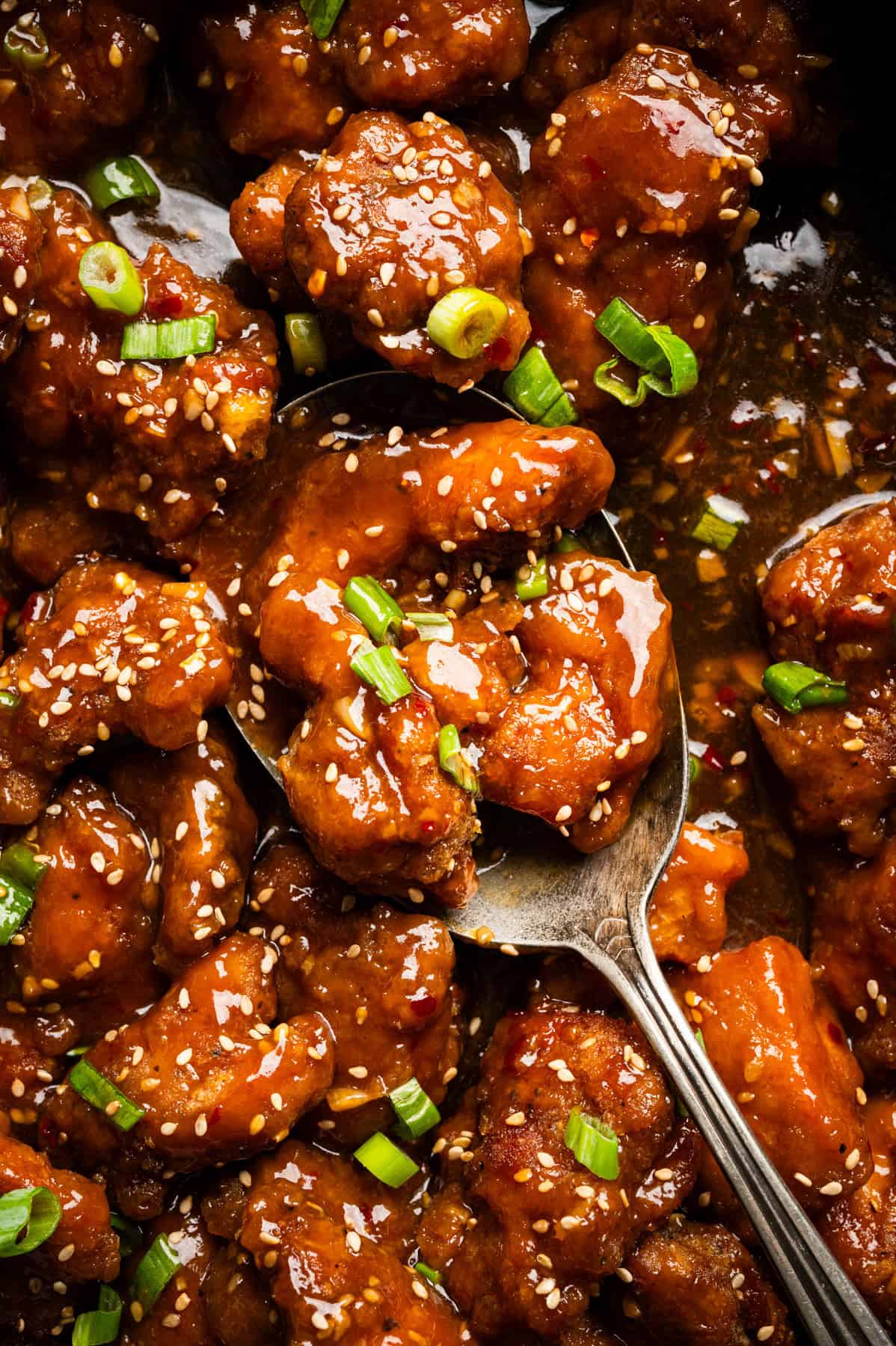 Orange Chicken Close-up of sticky, glazed chicken pieces garnished with sesame seeds and chopped green onions. A spoon scoops up some of the saucy chicken from the dish.