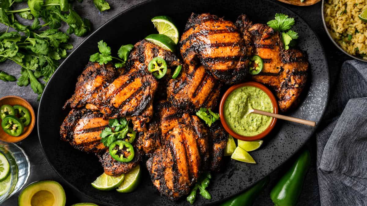 A black platter of grilled, marinated Peruvian chicken thighs garnished with cilantro, lime wedges, and sliced jalape&ntilde;os, served with a bowl of green sauce. 