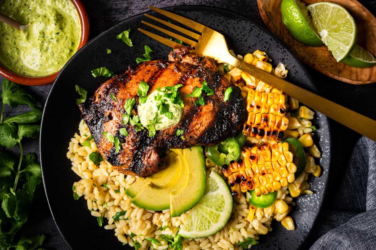 A black plate with Peruvian grilled chicken thigh topped with green sauce, served over rice with sliced avocado, lime wedges, grilled corn, and fresh herbs; a gold fork rests on the plate. Green sauce and lime are on the side.