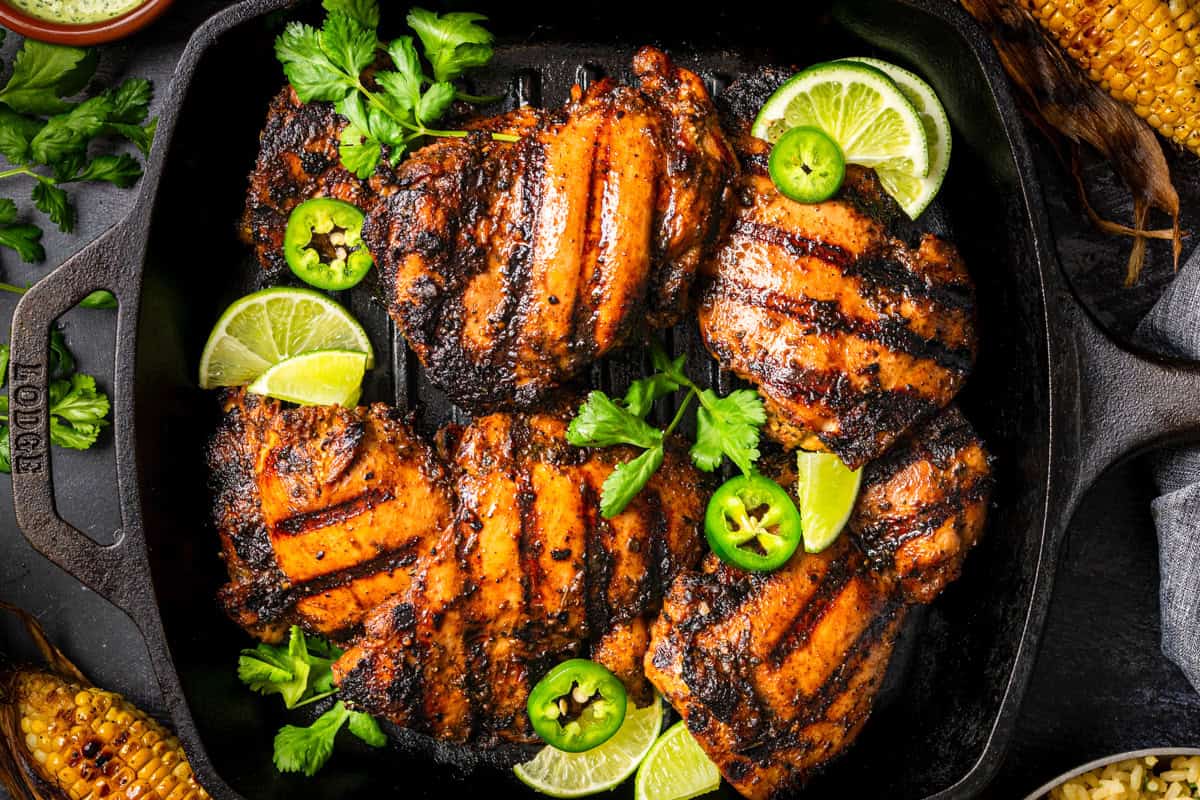 Four grilled chicken thighs with char marks on a grill pan, garnished with fresh cilantro, lime wedges, and sliced jalape&ntilde;os.