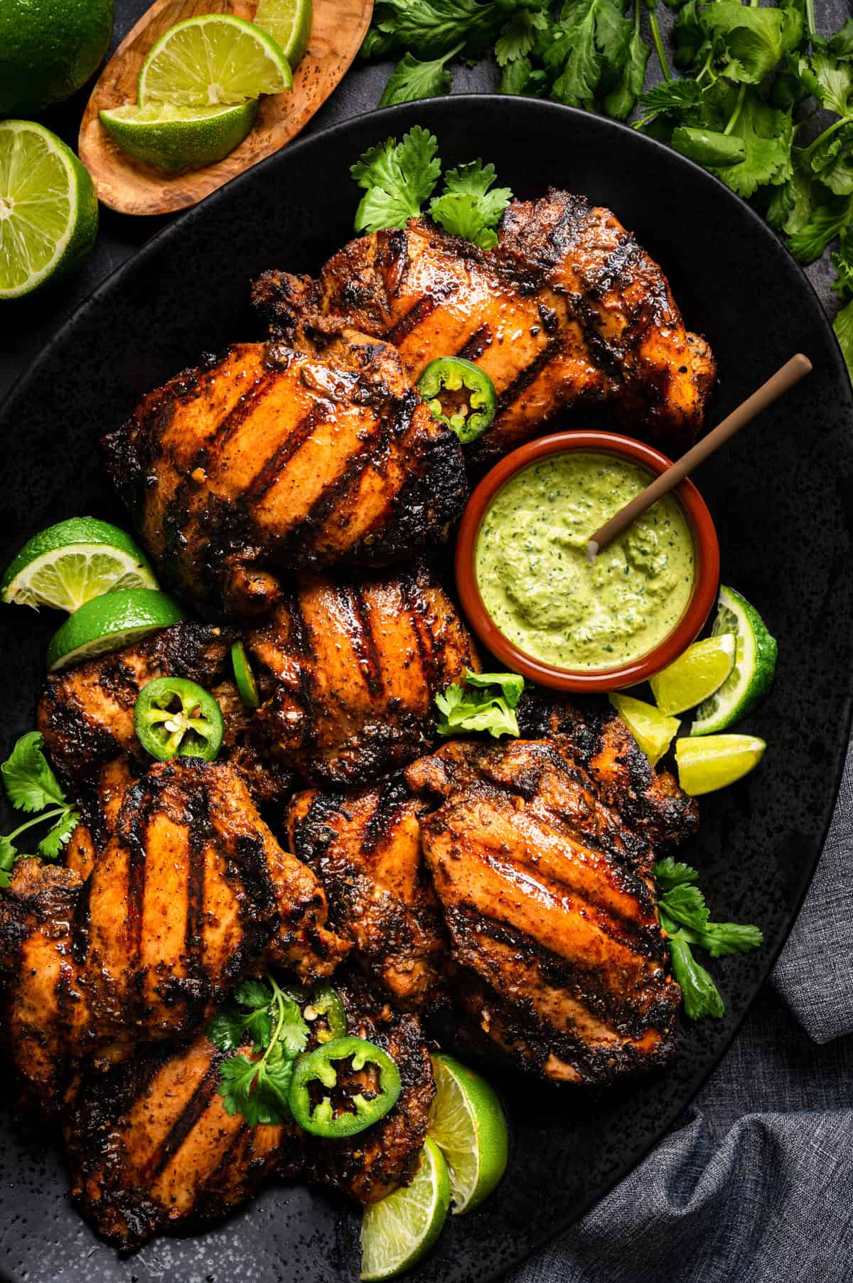 A black platter with Peruvian grilled chicken thighs garnished with lime wedges, cilantro, and jalape&ntilde;o slices, served with a small bowl of creamy green sauce and a spoon.