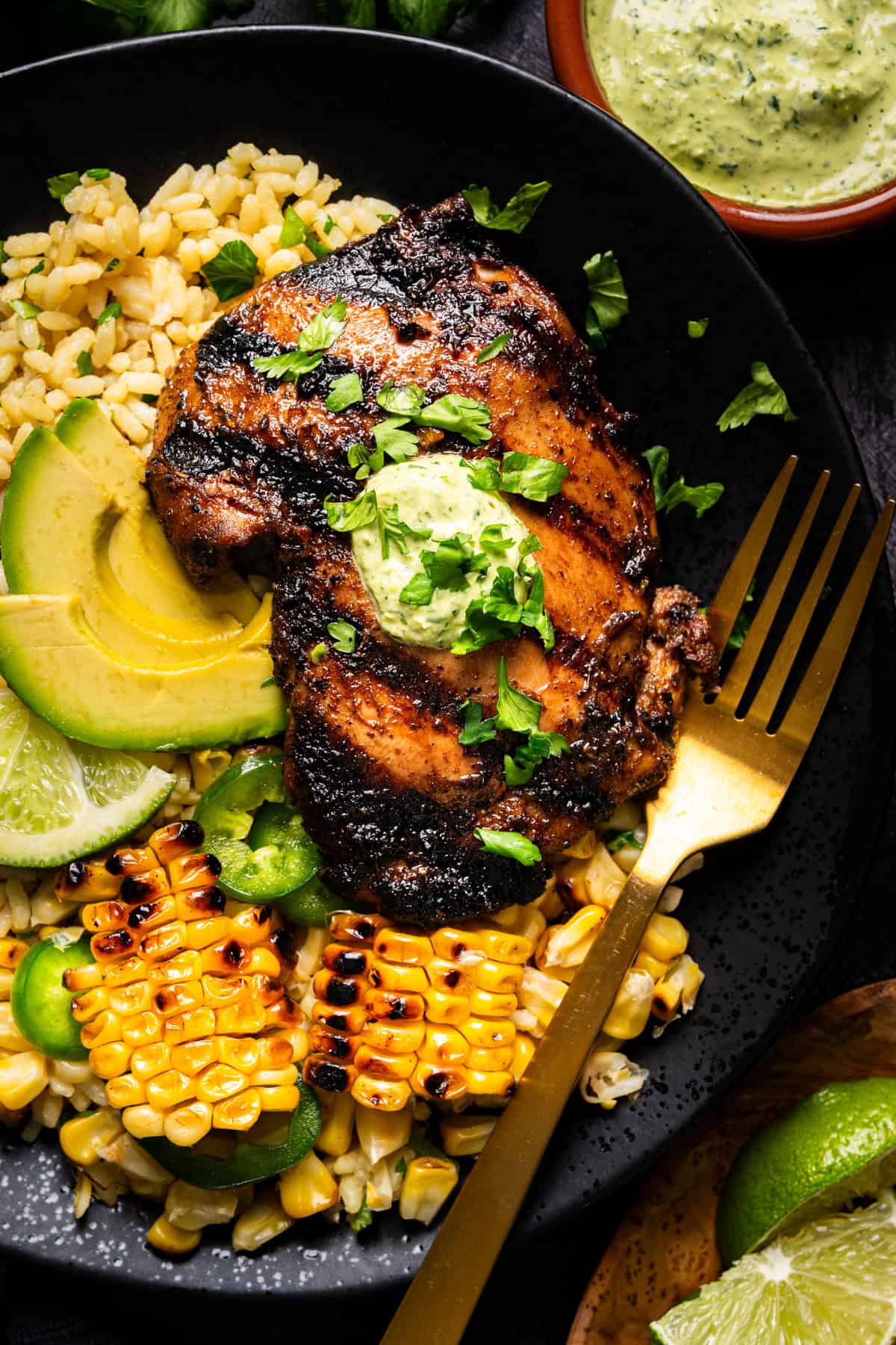 A black plate with Peruvian grilled chicken topped with green sauce, sliced avocado, lime wedges, grilled corn, rice, and jalape&ntilde;o slices. A gold fork rests on the plate, and a small bowl of green sauce is nearby.