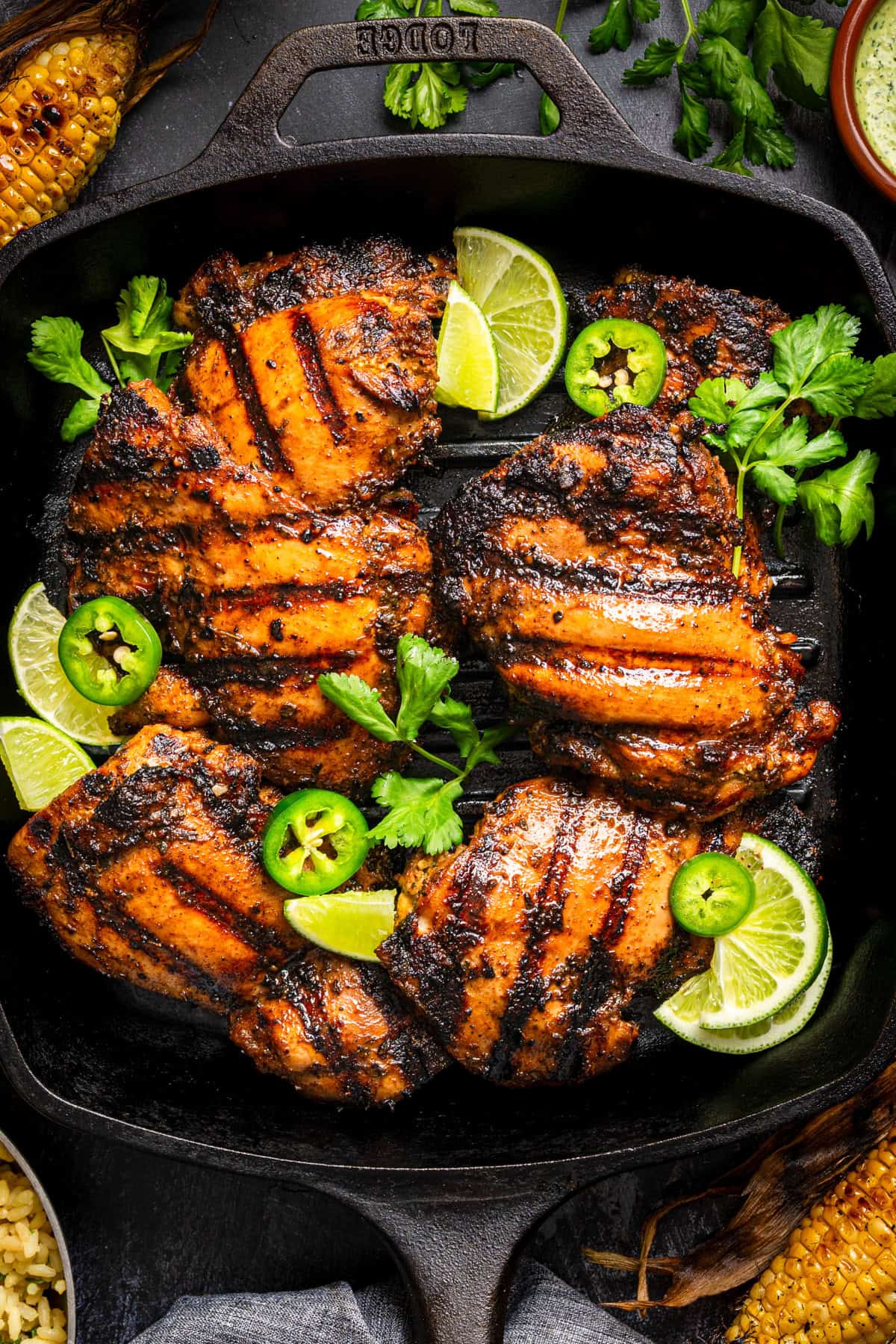 Peruvian Grilled chicken thighs with char marks in a cast iron pan, garnished with lime wedges, fresh cilantro, and sliced jalape&ntilde;os. Roasted corn and rice are partially visible around the pan.