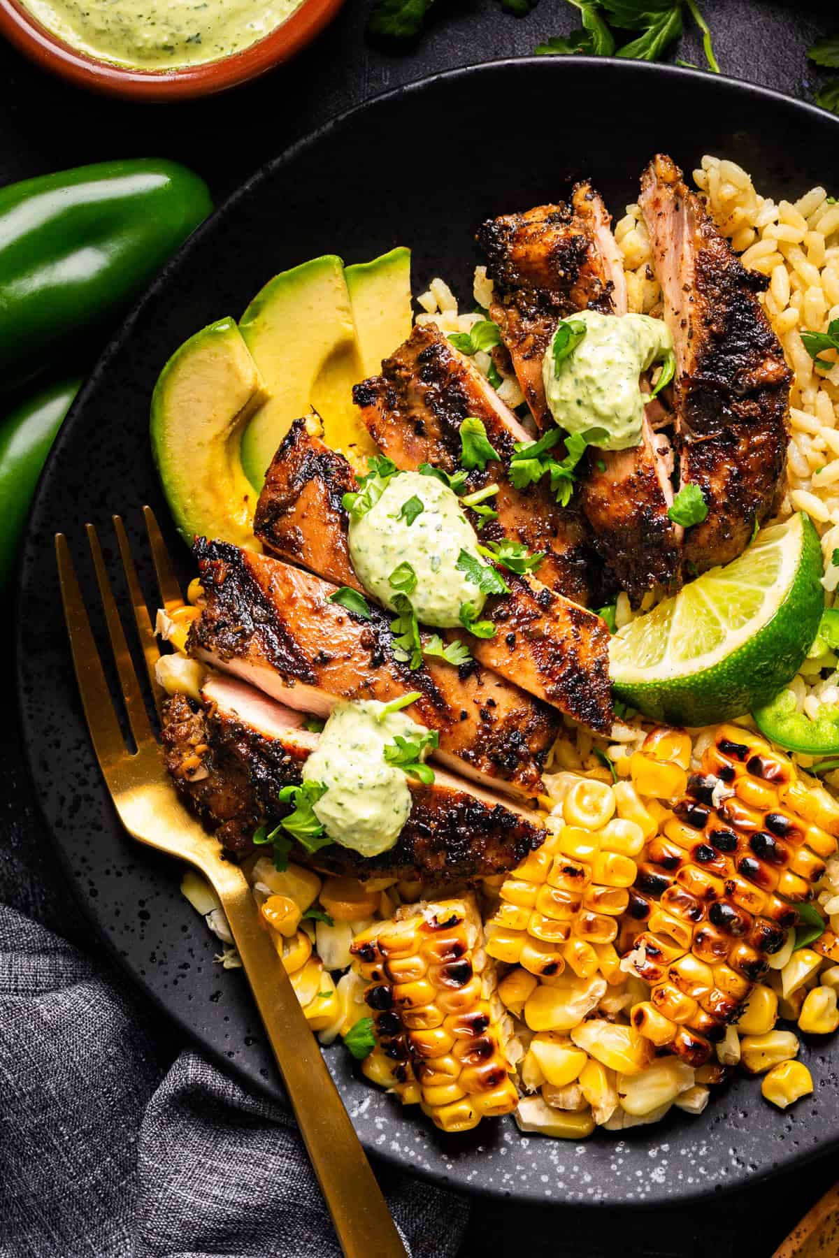 A black plate with grilled, sliced Peruvian chicken topped with green sauce, grilled corn, avocado slices, lime wedge, and rice, garnished with fresh herbs. 