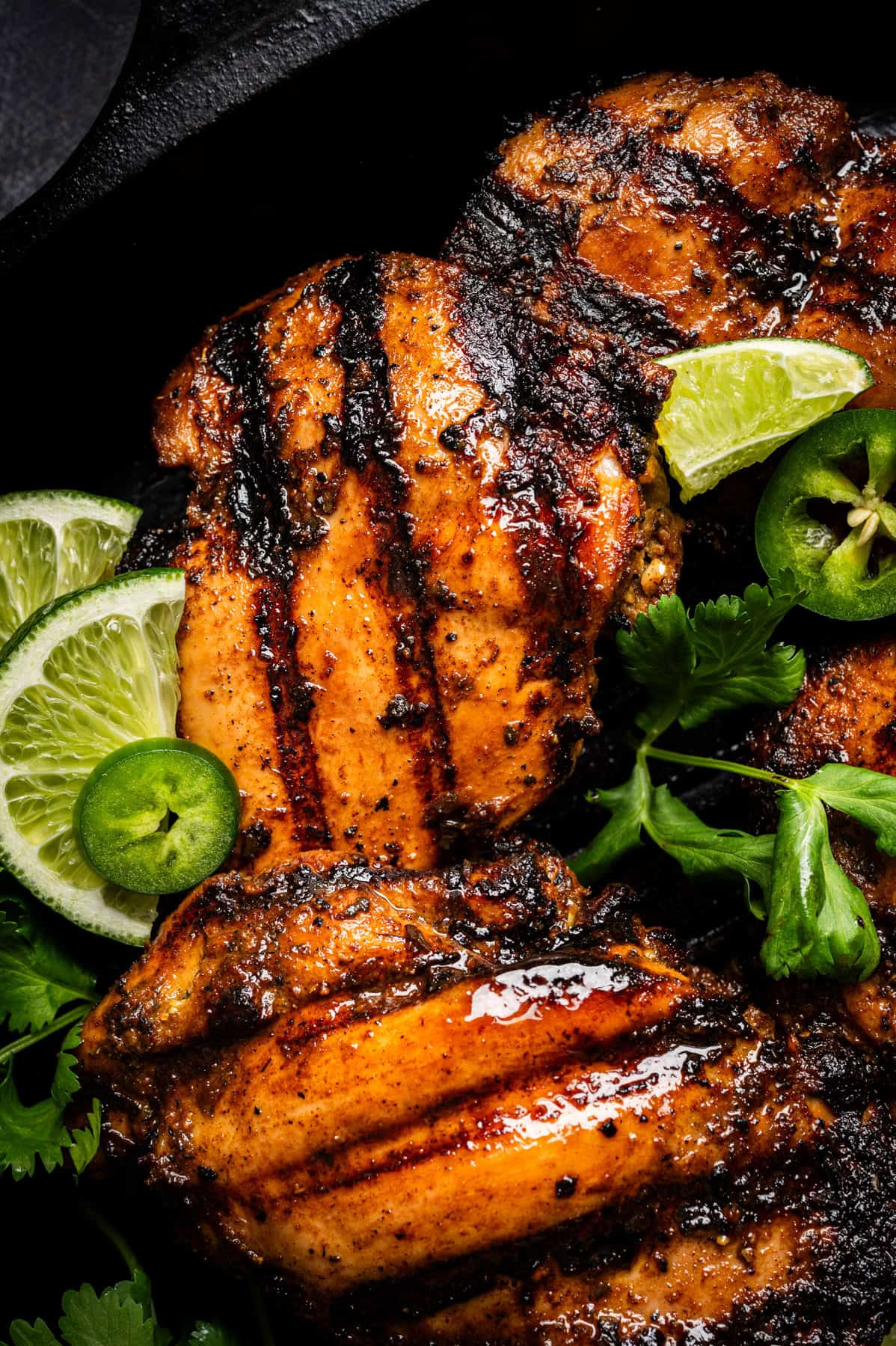Grilled, seasoned Peruvian chicken thighs with char marks, garnished with fresh lime slices, jalape&ntilde;o, and cilantro on a dark background.