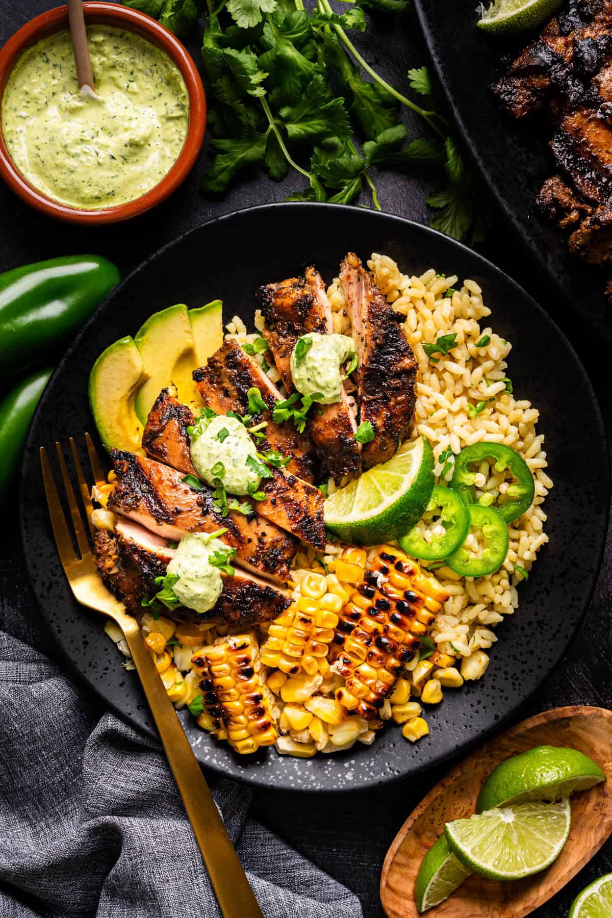 A black plate with grilled Peruvian chicken slices topped with green sauce, yellow rice, grilled corn, avocado slices, lime wedges, and jalape&ntilde;o slices, served with a gold fork. A bowl of creamy sauce and fresh herbs are nearby.