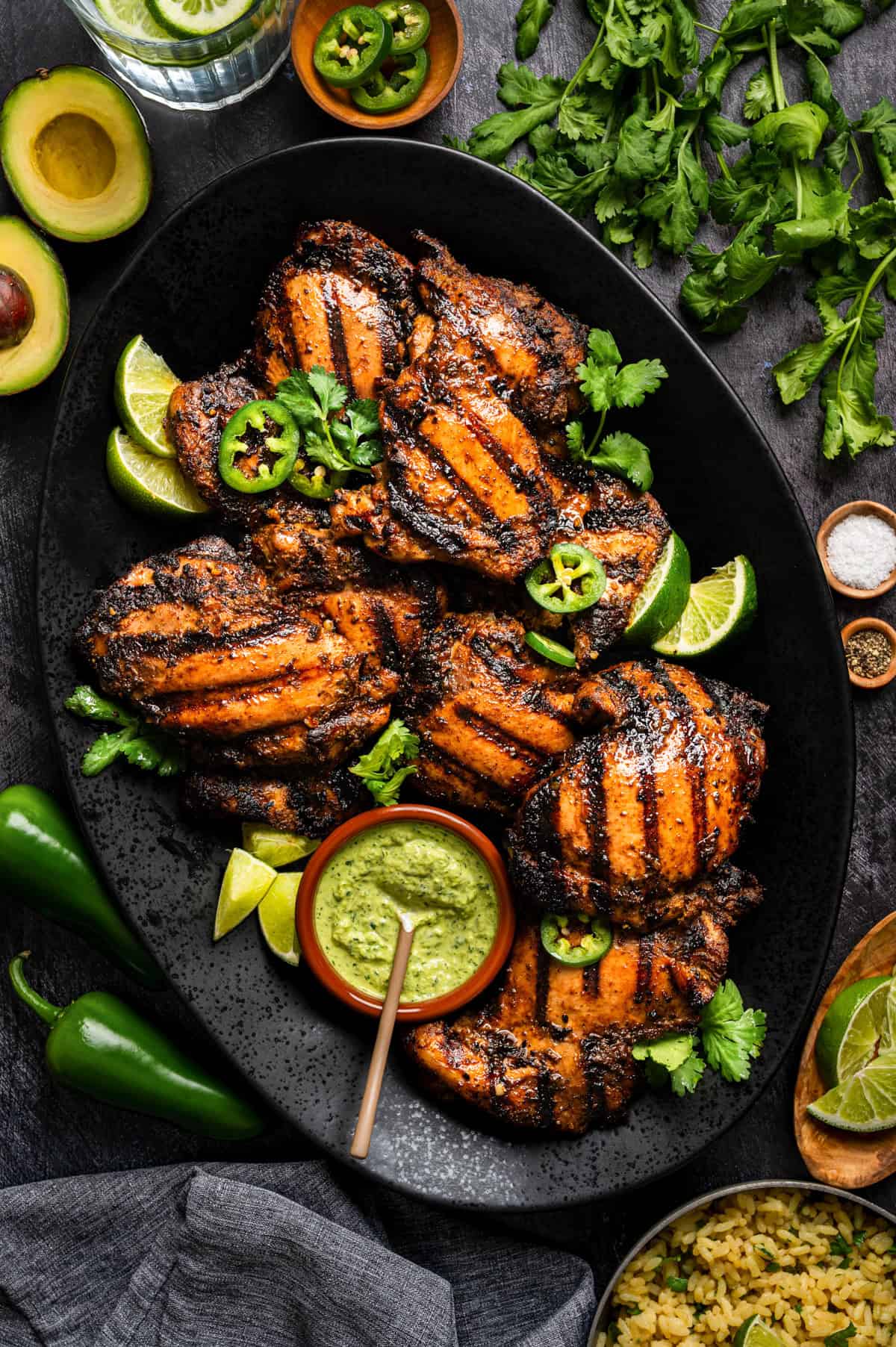 Peruvian Grilled chicken thighs with char marks on a black platter, garnished with lime wedges, cilantro, and sliced jalape&ntilde;os. Surrounded by avocado halves, fresh herbs, jalape&ntilde;os, rice, and a bowl of green sauce.