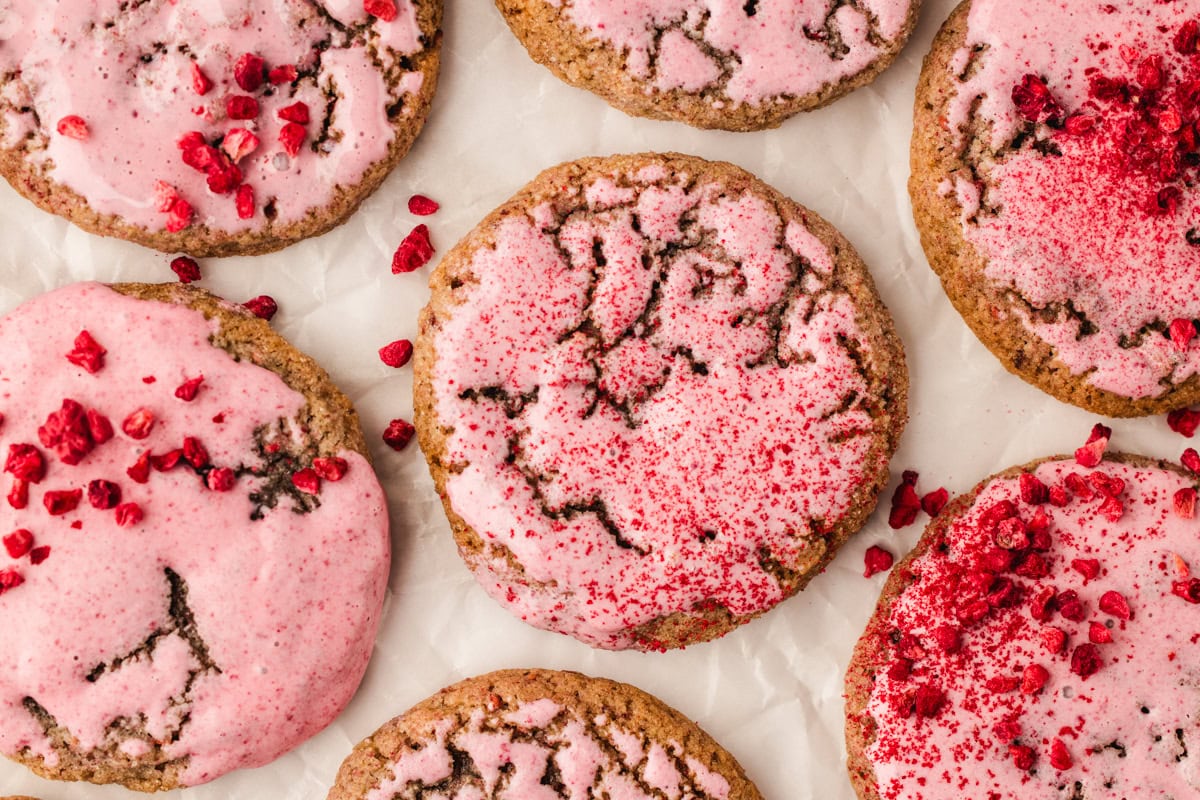 Six raspberry cookies with cracked tops are topped with pink icing and sprinkled with vibrant red berry pieces, arranged on white parchment paper.