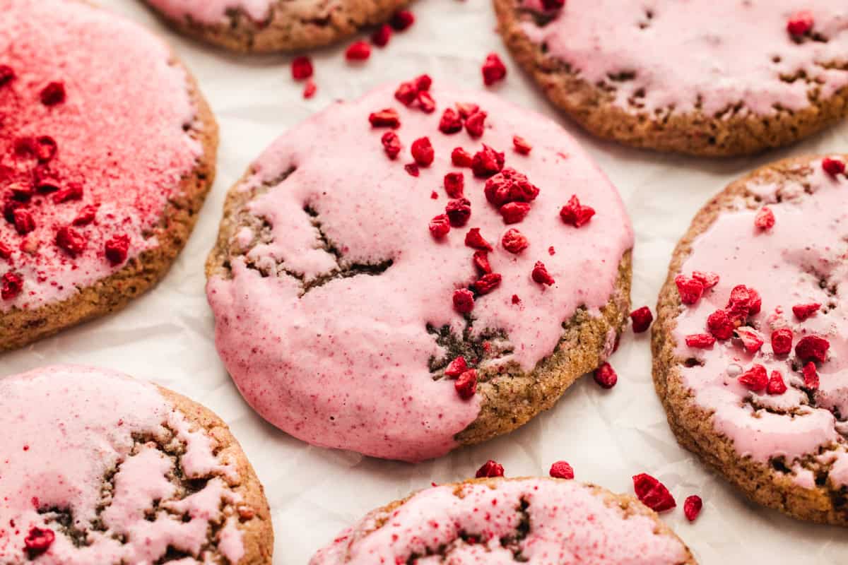 Several raspberry cookies with pink icing are arranged on parchment paper. Some cookies are partially covered in icing and sprinkled with small red pieces, while others have a full layer of pink icing on top.