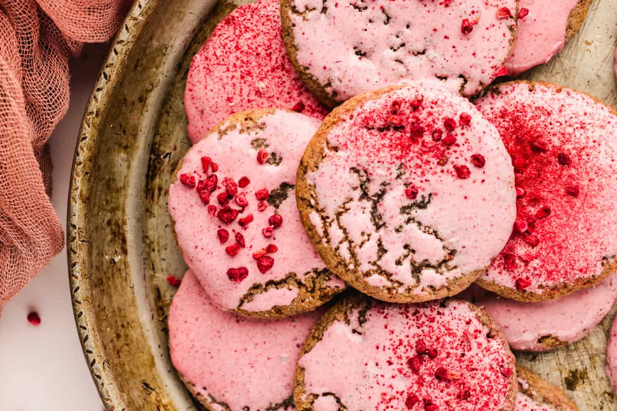 A close-up of round raspberry cookies with pink icing and red sprinkles, arranged on a rustic metal tray.