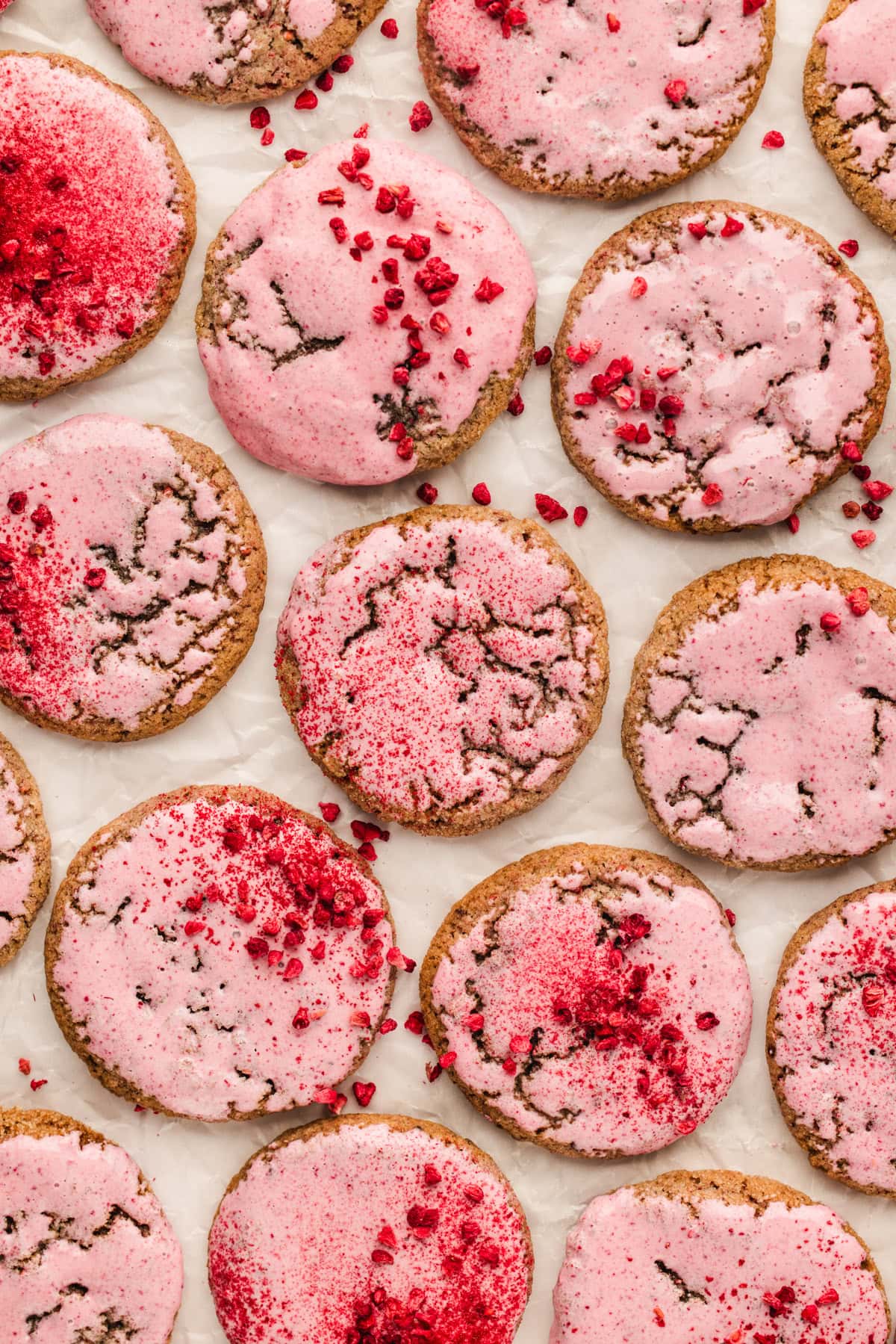 Several raspberry cookies with a cracked surface are topped with pink icing and sprinkled with red crumbs, arranged in a grid pattern on white parchment paper.