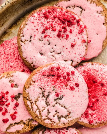 A close-up of several round raspberry cookies with cracked surfaces, covered in a pink icing and sprinkled with red raspberry bits, arranged on a metal tray.