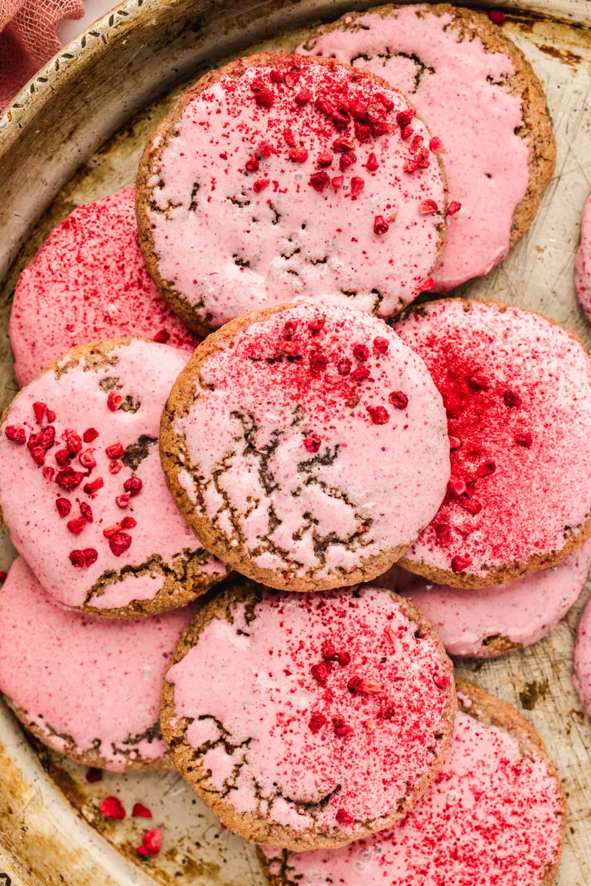A tray of round raspberry cookies topped with pink icing and sprinkled with crushed dried raspberries. The cookies have a crackled surface and are arranged in a slightly overlapping pattern.