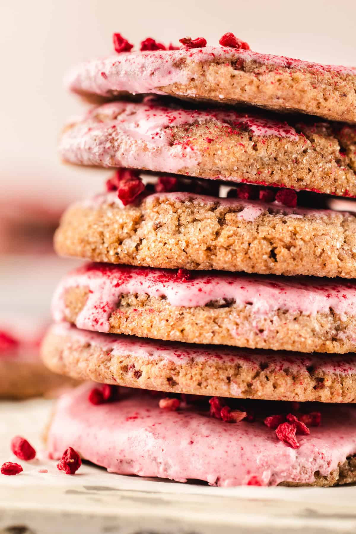 A close-up of a stack of raspberry cookies topped with pink glaze and sprinkled with pieces of dried raspberries on top, set on a light surface.