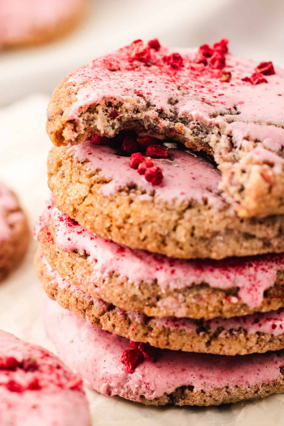 A stack of raspberry cookies topped with pink frosting and sprinkled with red berry pieces, with the top cookie partially bitten.