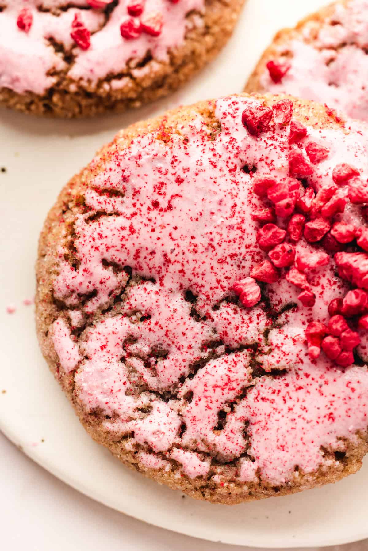 A close-up of a raspberry cookie topped with pink icing and sprinkled with dried raspberry pieces, on a white plate. Another similar cookie is partially visible in the background.