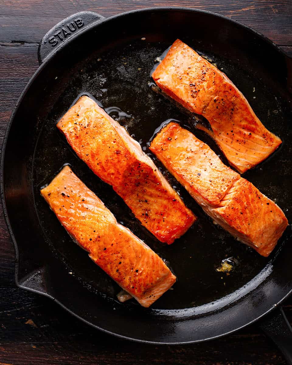 Four seared salmon fillets cooking in a black cast iron skillet, with golden-brown crispy edges.
