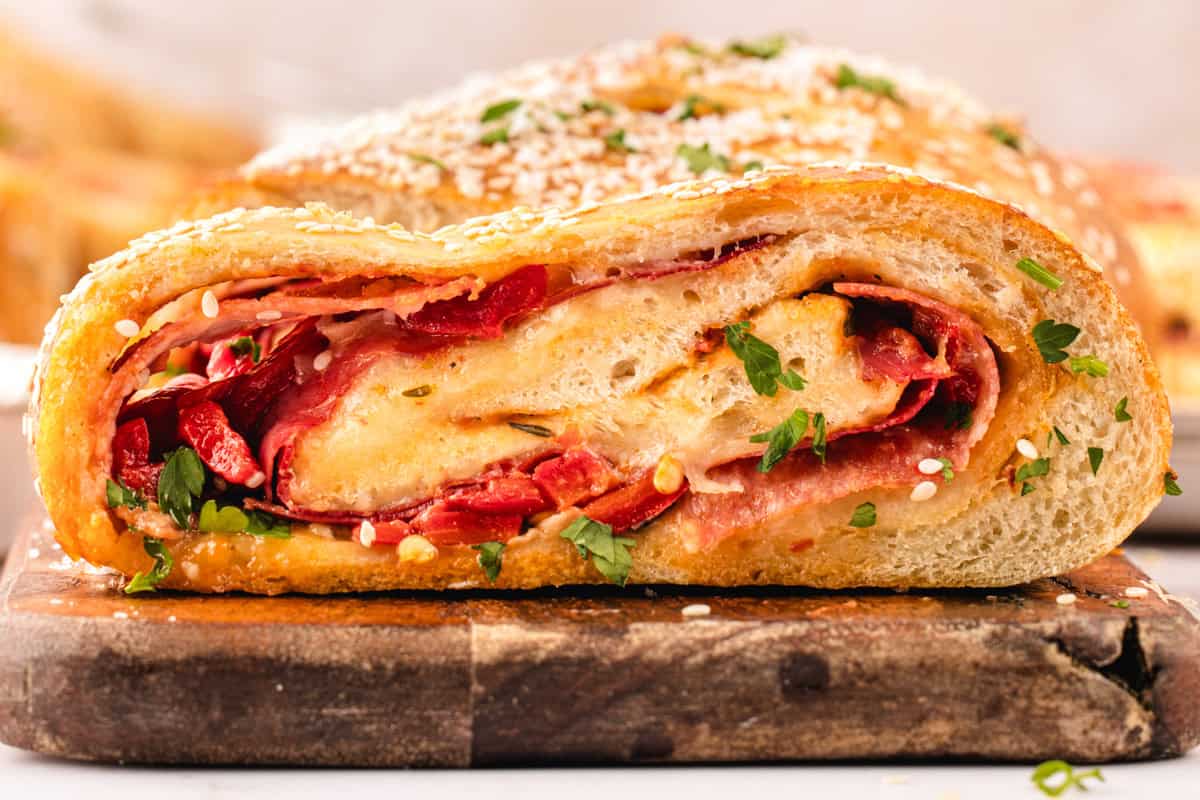 Stromboli A close-up of a sliced stromboli filled with melted cheese, meat, red peppers, and fresh herbs, all wrapped in golden baked dough and topped with sesame seeds.