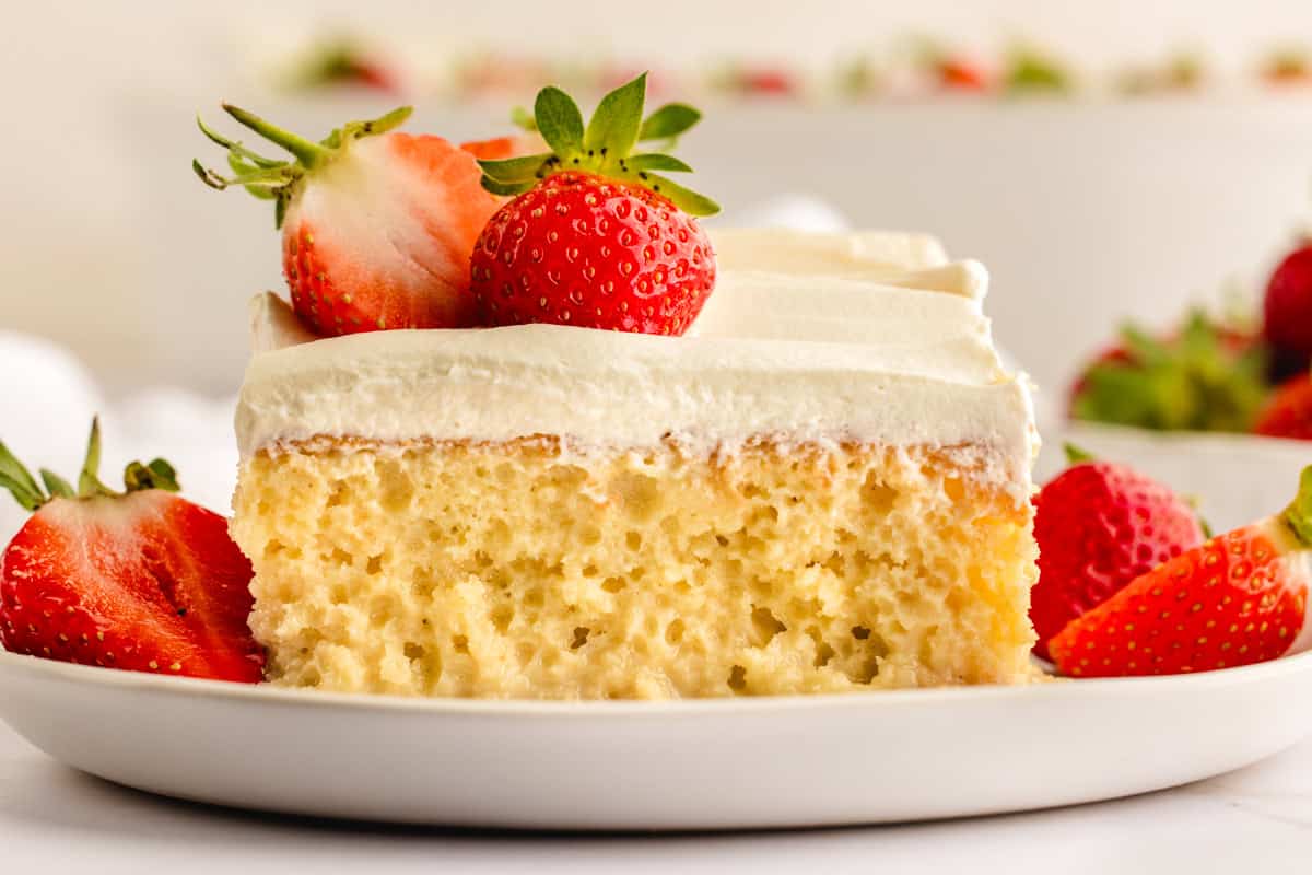 A slice of moist tres leches cake topped with whipped cream and a fresh strawberry, served on a white plate with more strawberries around it.