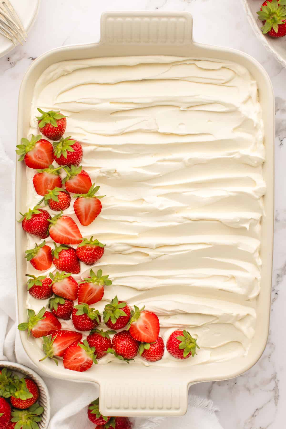 A rectangular baking dish filled with smooth, white whipped cream and decorated with a cluster of fresh, halved strawberries and green leaves in one corner.