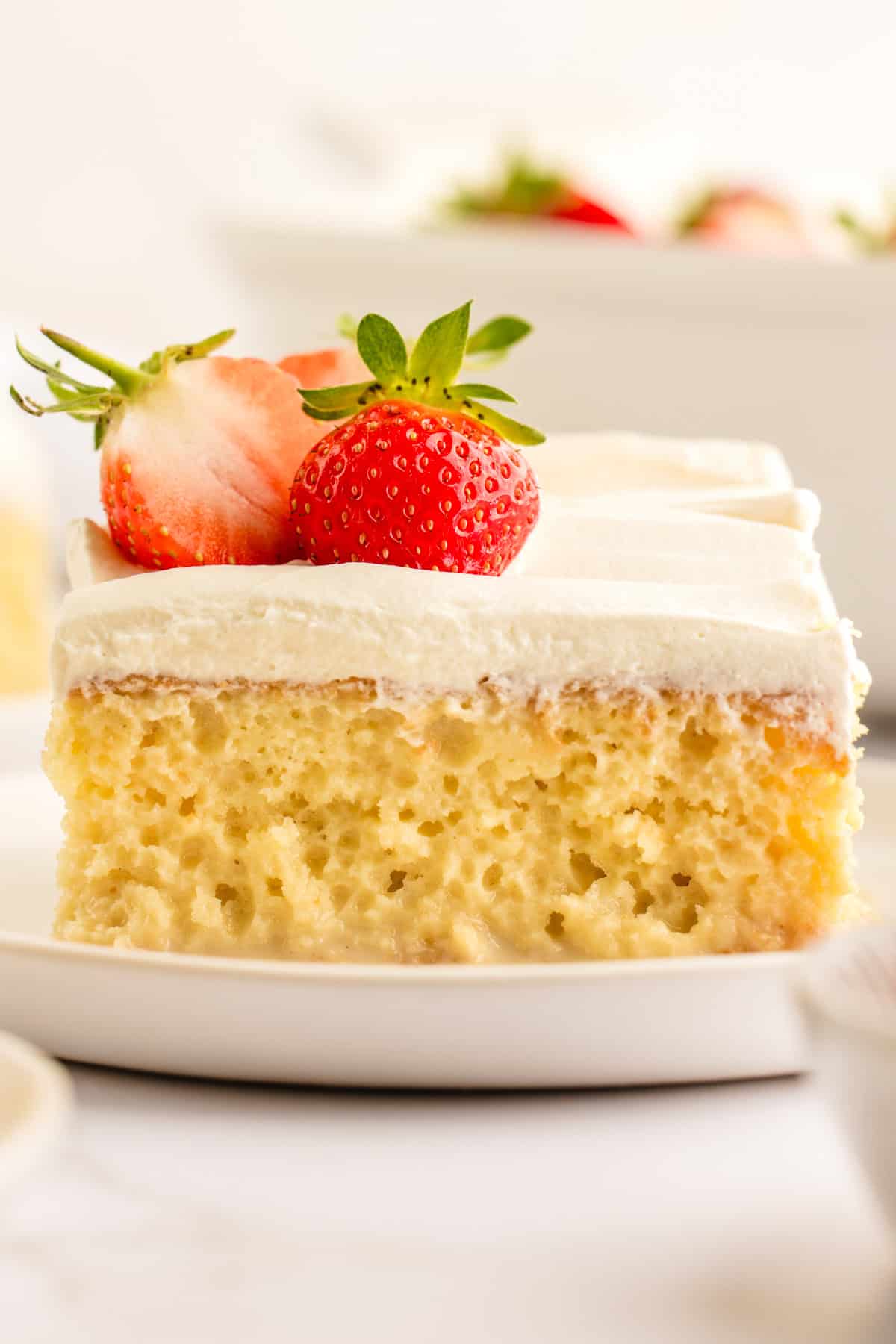 A slice of tres leches cake topped with whipped cream and two fresh strawberries sits on a white plate. The cake appears moist and spongy with a creamy topping.