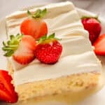 A slice of tres leches cake topped with whipped cream and fresh strawberries, set on a white plate with sliced strawberries on the side.