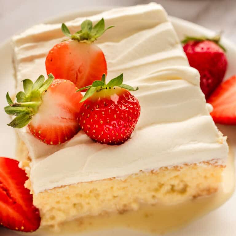A slice of tres leches cake topped with whipped cream and fresh strawberries, set on a white plate with sliced strawberries on the side.