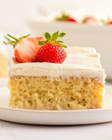 A slice of moist tres leches cake topped with white whipped cream and a halved strawberry, served on a white plate.