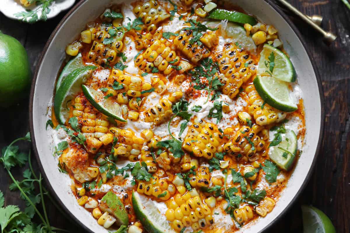 A white bowl filled with Mexican street corn dip, featuring grilled corn kernels, creamy sauce, lime wedges, chopped cilantro, and a sprinkle of chili powder. 