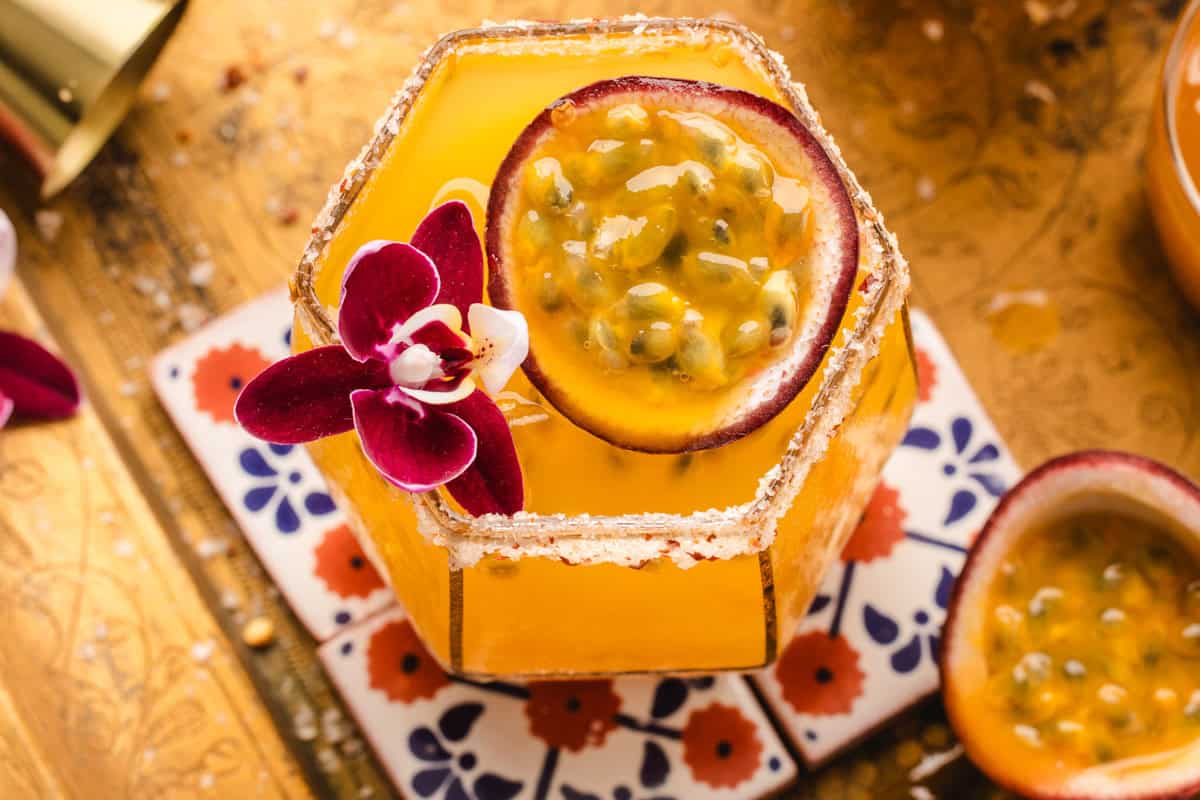 A vibrant yellow passion fruit margarita in a hexagonal glass, garnished with a halved passion fruit and a purple orchid, sits on a colorful tile coaster atop a gold patterned surface. The rim of the glass is coated with sugar.