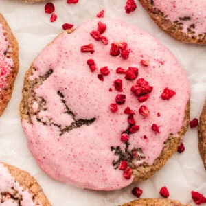A close-up of a cookie with pink icing and small pieces of dried red berries on top, placed on white parchment paper.