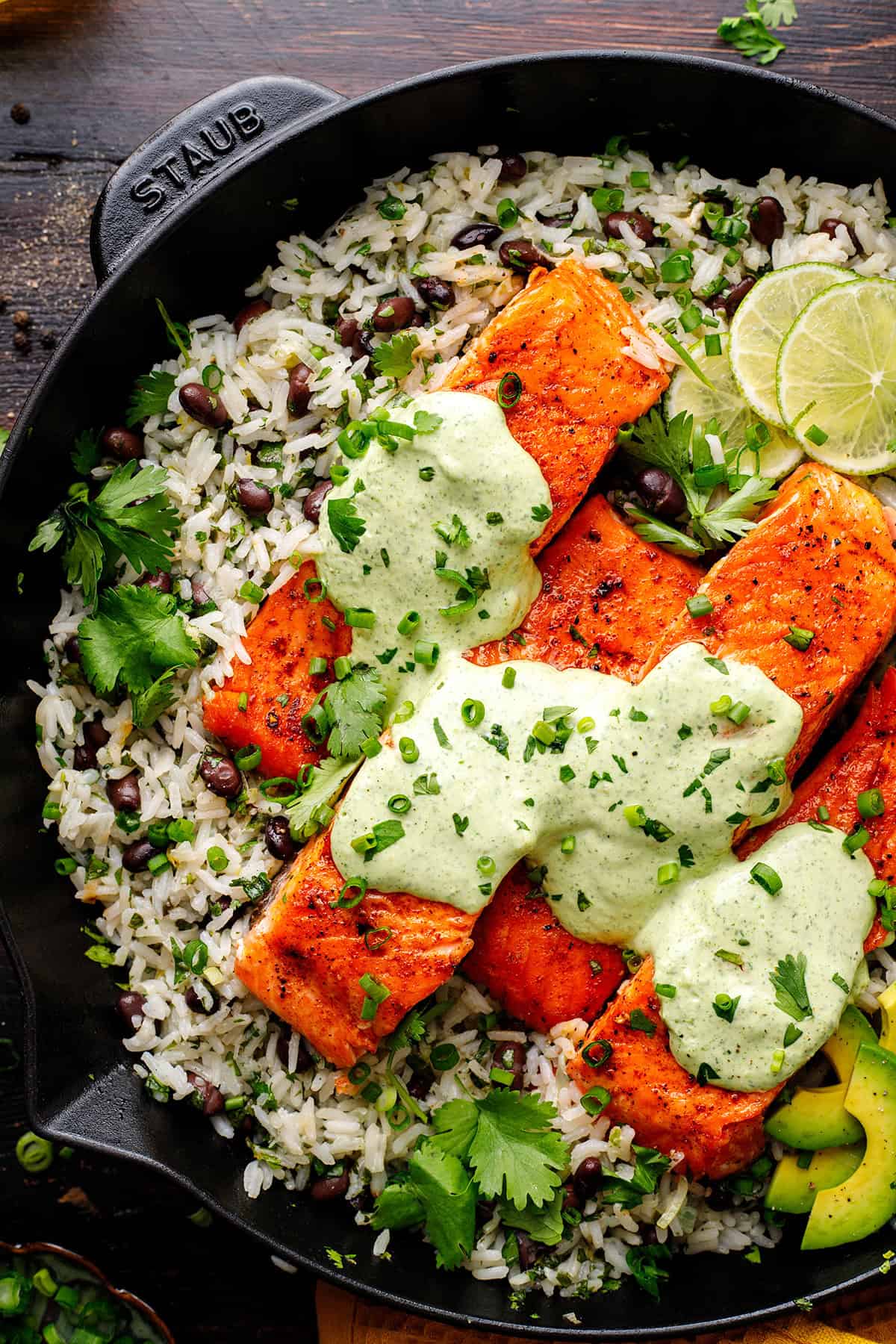 A cast-iron skillet filled with rice, black beans, and herbs, topped with three pieces of cooked salmon drizzled with a creamy green sauce. Garnished with lime slices and fresh cilantro.