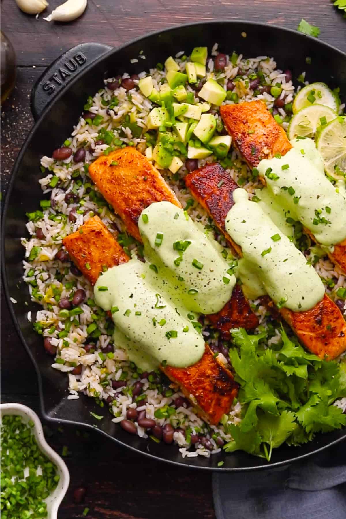A cast-iron skillet with three pieces of seasoned salmon on a bed of rice, black beans, and avocado, topped with green cilantro-lime creamy sauce and garnished with lime and fresh cilantro.