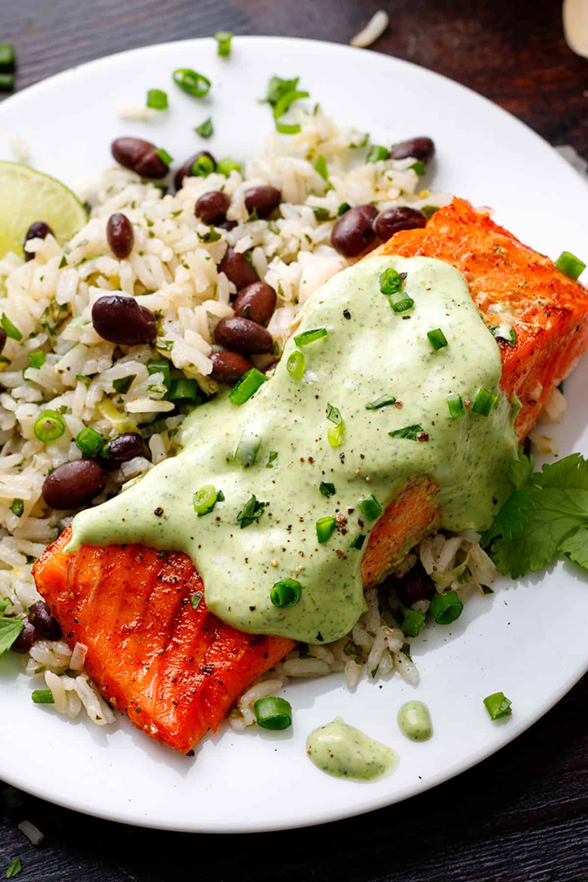 A plate with pan-seared salmon topped with green creamy cilantro-lime sauce, served alongside rice mixed with black beans, cilantro, and herbs. A lime wedge and cilantro garnish the dish.