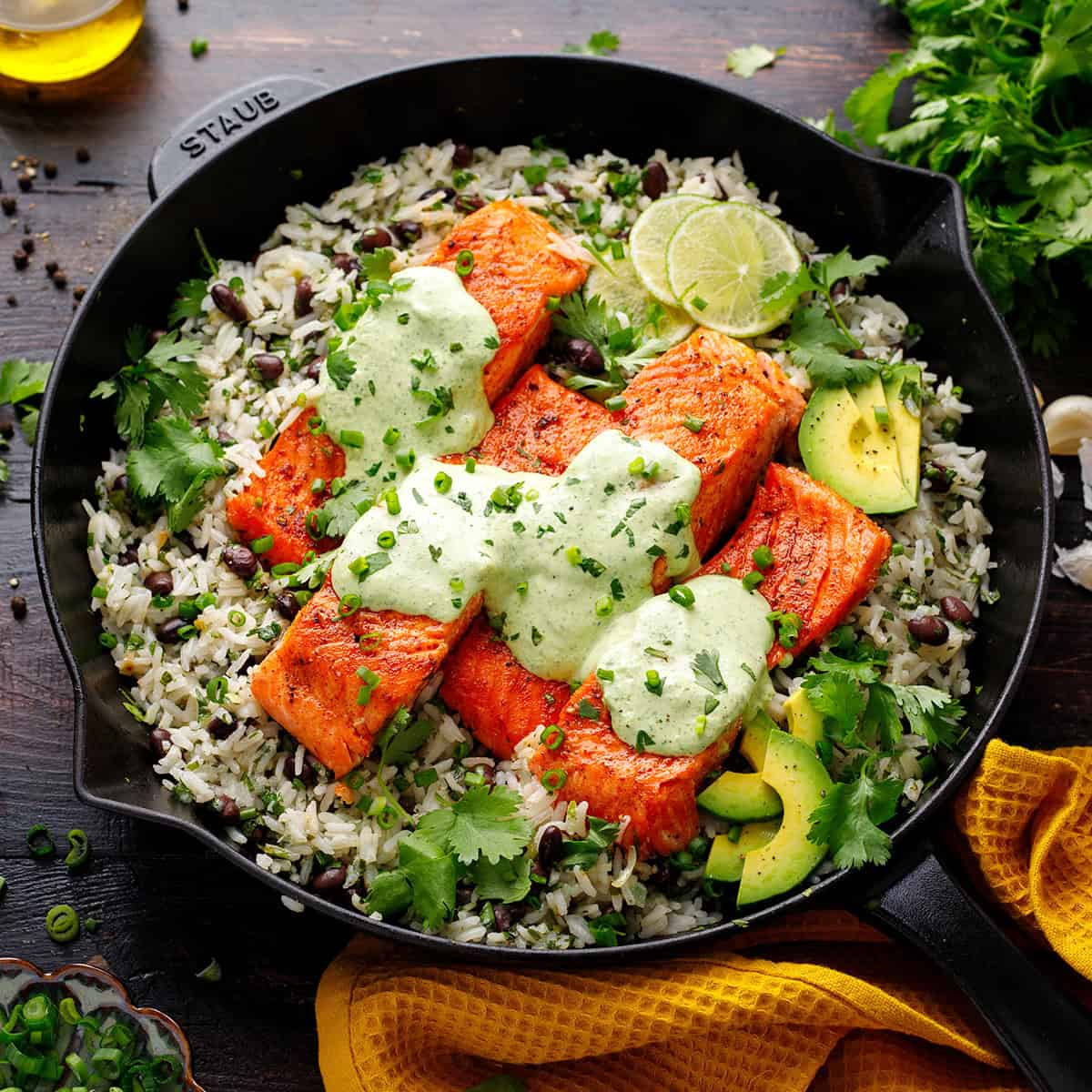 A cast-iron skillet with seared salmon fillets on cilantro rice with black beans, avocado slices, lime wedges, and green sauce, garnished with fresh cilantro and chopped chives.