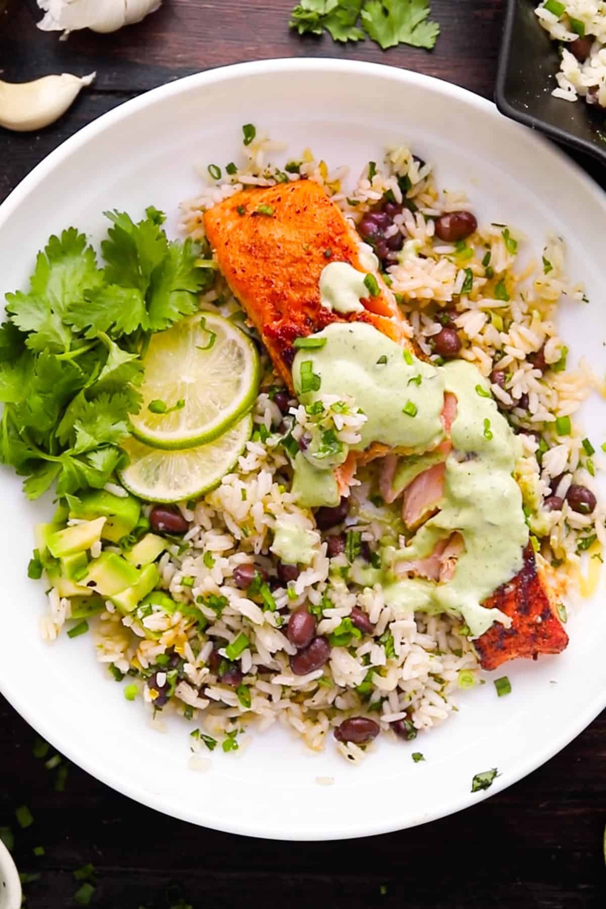 A plate of rice mixed with black beans and herbs, topped with a piece of pan-seared salmon drizzled with creamy cilantro-lime sauce. Garnished with avocado chunks, lime slices, and fresh cilantro.