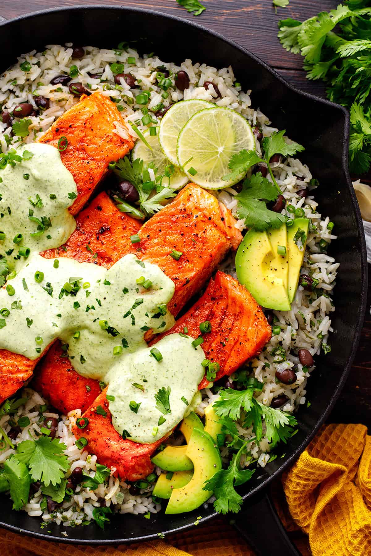 A cast-iron skillet filled with pan-seared salmon fillets on a bed of rice and black beans, topped with creamy green cilantro-lime sauce, sliced avocado, lime wedges, and fresh cilantro, garnished with chopped chives.