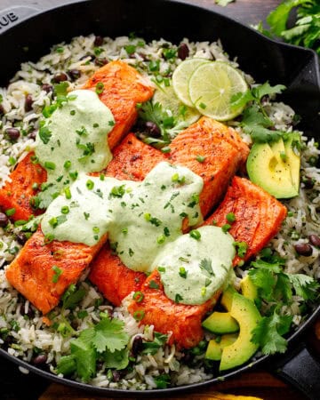 A cast iron skillet filled with pan-seared salmon fillets on a bed of cilantro rice and black beans, topped with creamy green cilantro-lime sauce, avocado slices, lime, and fresh herbs.