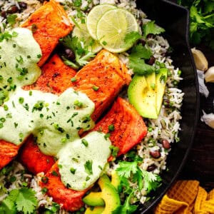 Grilled salmon fillets topped with creamy green cilantro-lime sauce, served on rice with black beans, avocado slices, fresh cilantro, lime slices, and chopped green onions in a black cast iron skillet.